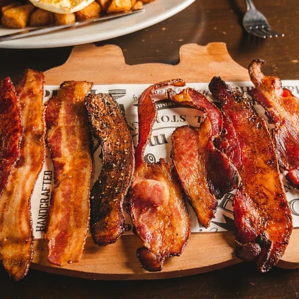 Crispy bacon strips on a wooden board with a breakfast plate in the background.