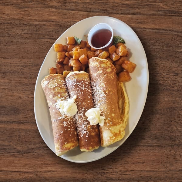 Three rolled pancakes with powdered sugar and whipped cream, roasted potatoes, and syrup on a white plate.